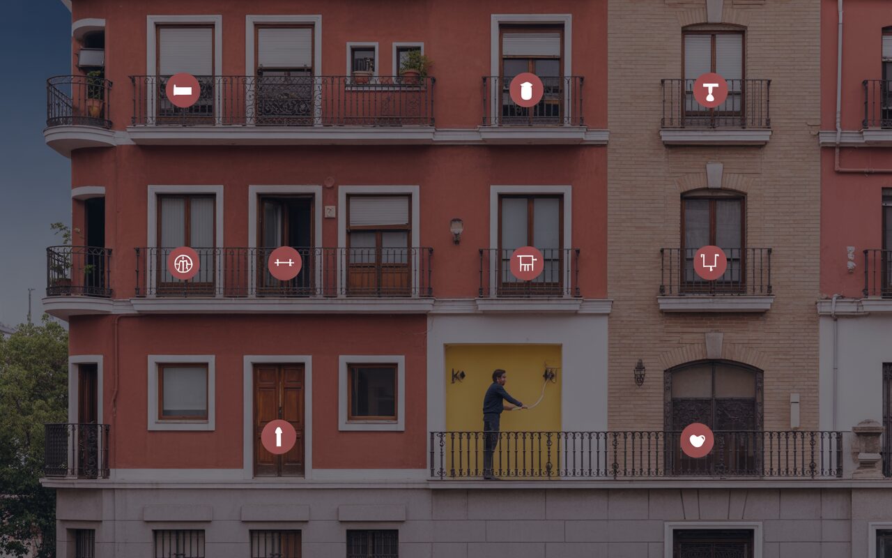 La fachada de un edificio residencial típico de Madrid, donde varios iconos de "hogar inteligente" (domótica) están superpuestos sobre los diferentes balcones y ventanas, y un hombre usa un rodillo de pintura en una pared.